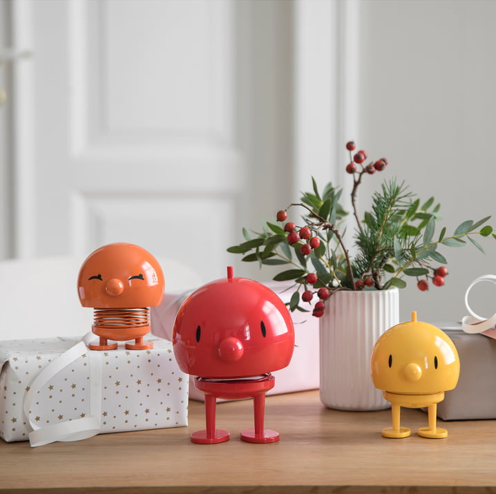Drei bunte Hoptimist-Figuren in Orange, Rot und Gelb auf einem Holztisch, umgeben von Geschenkboxen und einer Vase mit Zweigen und roten Beeren