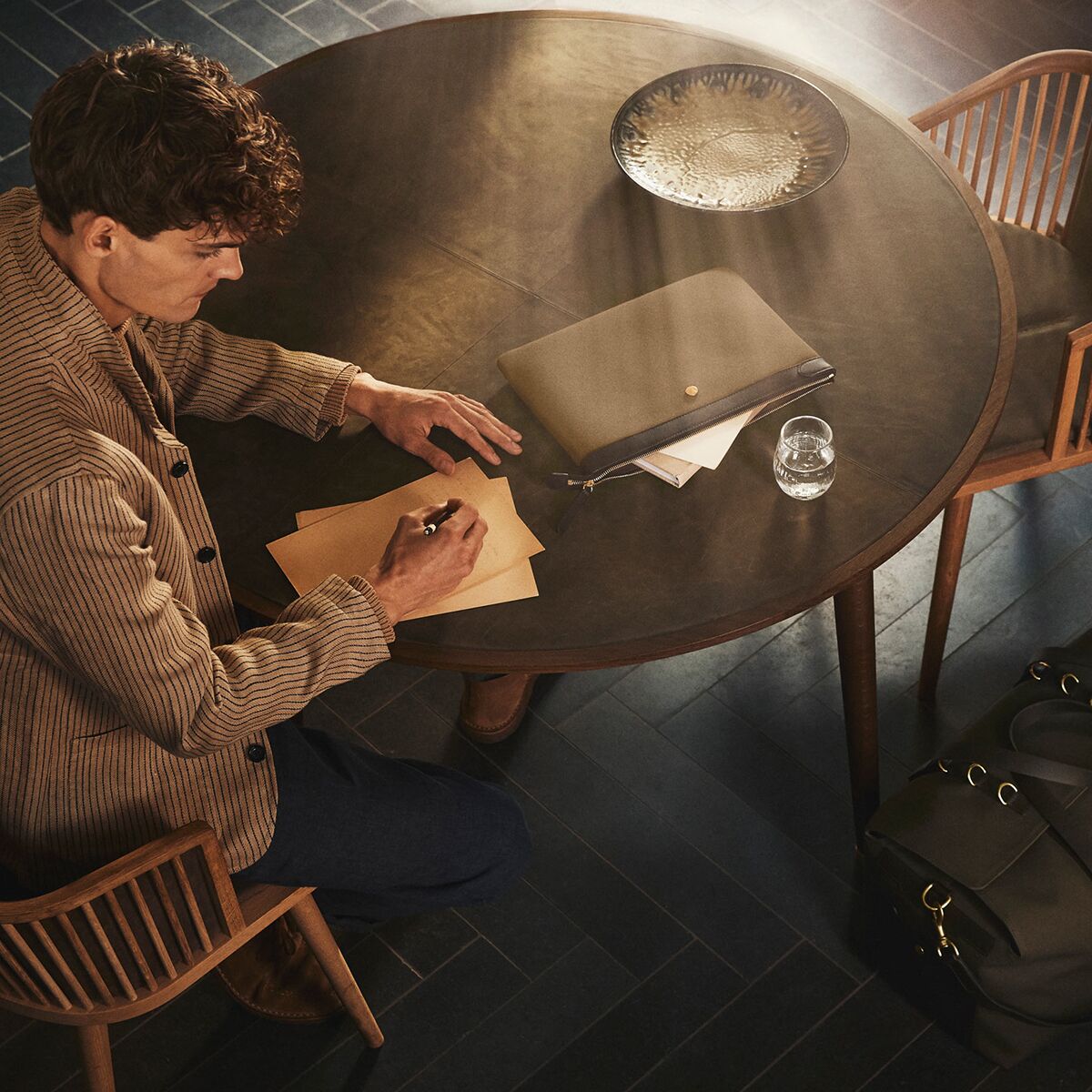 Ein Mann sitzt an einem runden Holztisch mit dunkler Oberfläche und schreibt mit einem Stift auf beiges Papier. Auf dem Tisch liegen eine elegante oliv-braune MISMO Tasche »POUCH LARGE«, ein silberfarbenes Tablett sowie Dokumente und ein Glas Wasser. Die Szene wirkt stilvoll und ruhig.