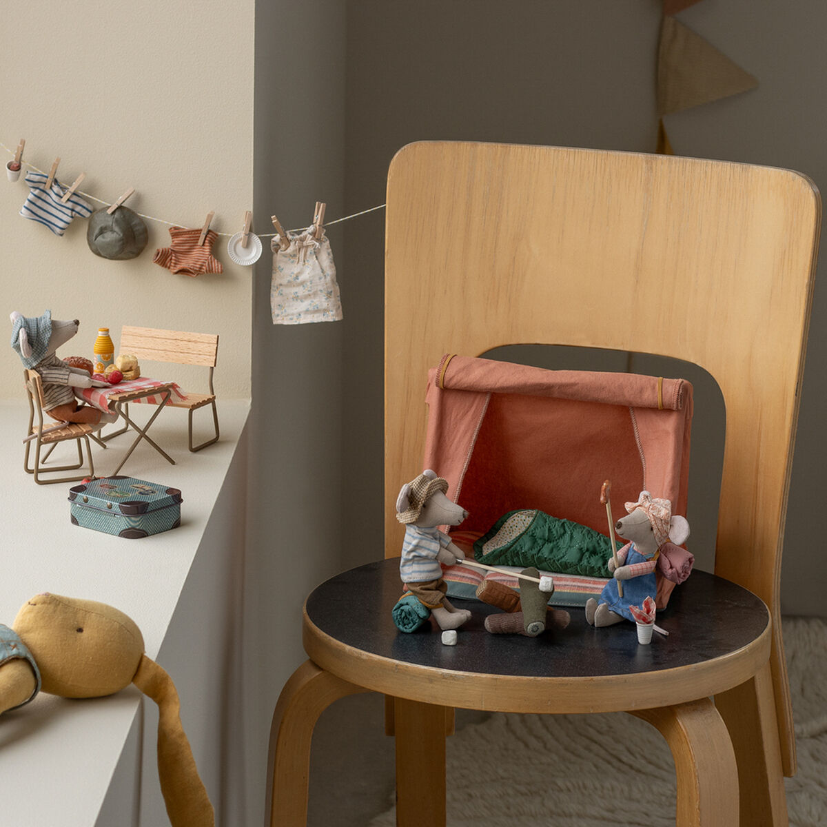Zwei Stoffmäuse mit Campingzubehör und einem kleinen Zelt auf einem Holzstuhl. Im Hintergrund sind Miniaturkleidung an einer Wäscheleine, Möbel und Dekorationen in sanften Farben im Kinderzimmer zu sehen.