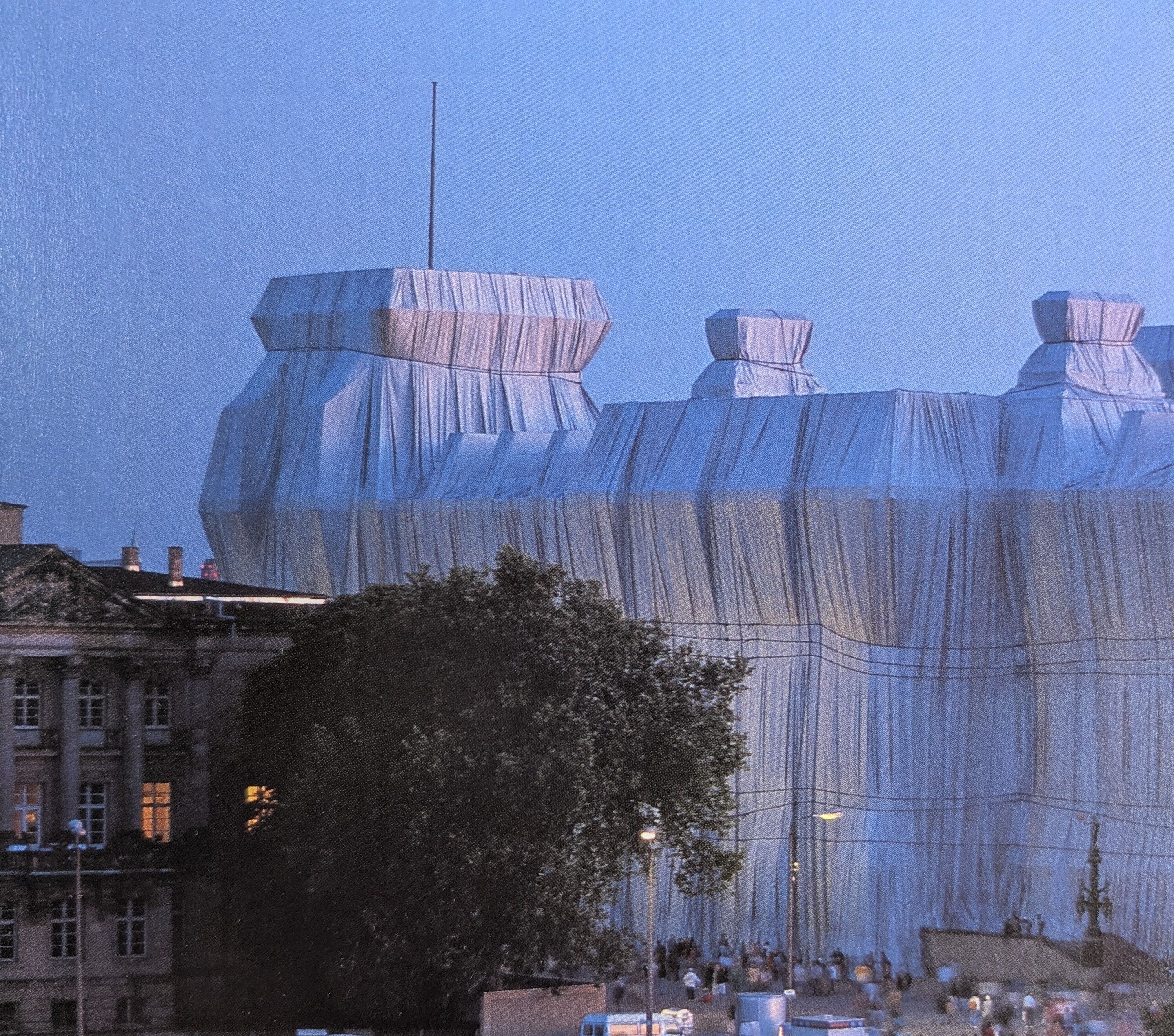 Foto des reichstaggebäudes in Berlin, das von Christo verhüllt wurde. Das Gebäude ist mit einem grauen Stoff bedeckt, Bäume sind im Vordergrund zu sehen.