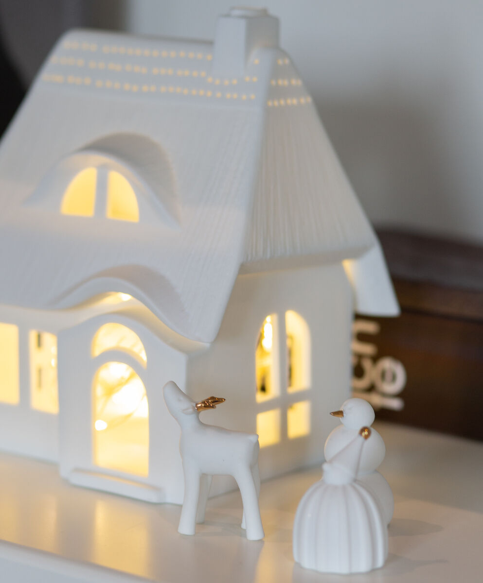 Ein weißes Porzellan-Lichthaus mit Reetdach steht auf einer Oberfläche. Im Inneren des Hauses leuchtet warmes Licht. Davor sind eine kleine Hirschfigur und eine Schneemannfigur aus Porzellan aufgestellt. Die Szene strahlt winterliche Gemütlichkeit aus und eignet sich als festliche Dekoration.