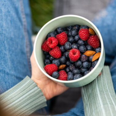 Person hält eine grüne Lunchbox mit Blaubeeren, Himbeeren und Mandeln, die auf ihrem Schoß sitzt.