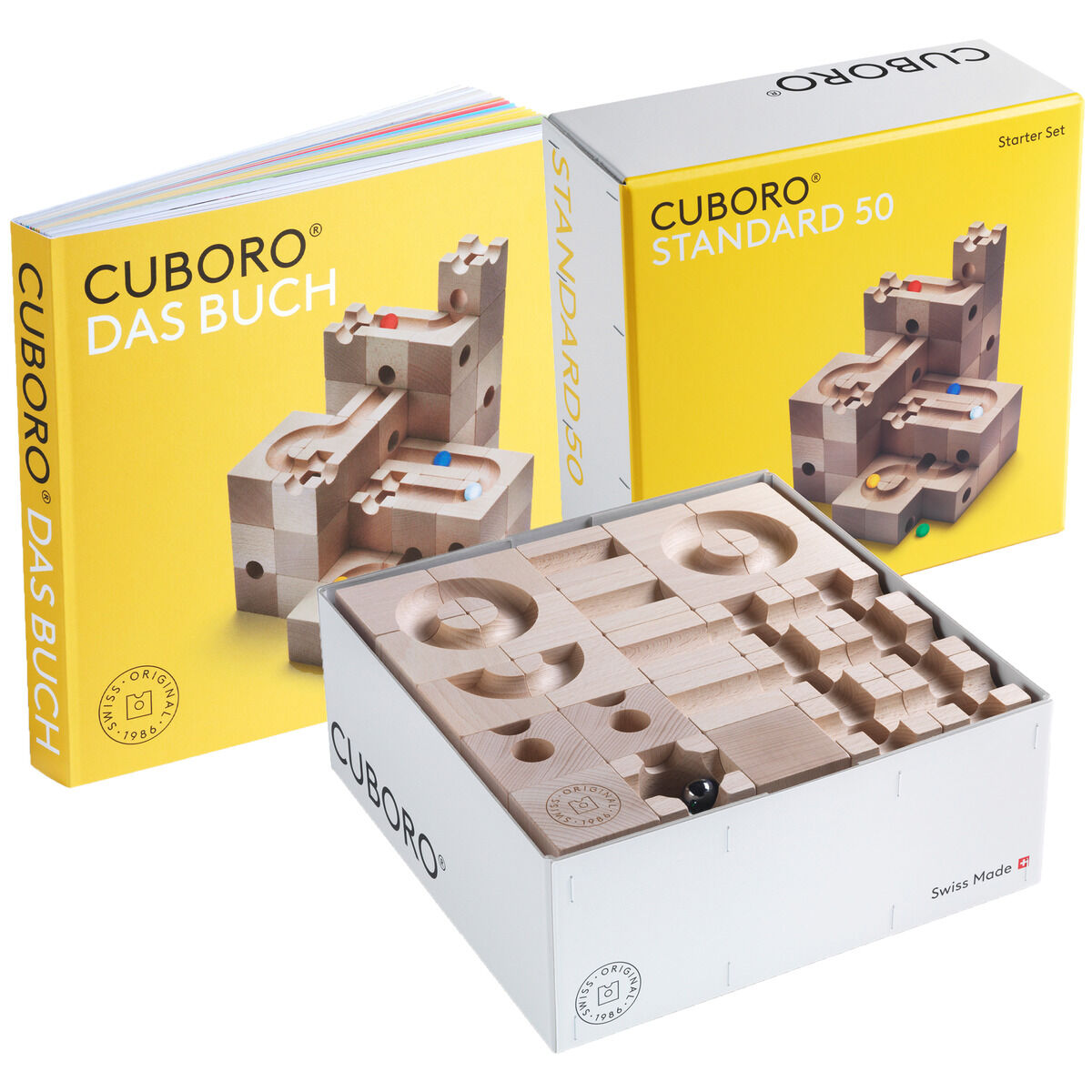 Das Bild zeigt das Kugelbahn-Set CUBORO STANDARD 50 sowie das Buch CUBORO DAS BUCH. Beide sind aus hellem Holz gefertigt und in gelben und weißen Verpackungen präsentiert. Die Boxen enthalten verschiedene Holzbausteine und Bahnen für den Bau einer Murmelbahn.