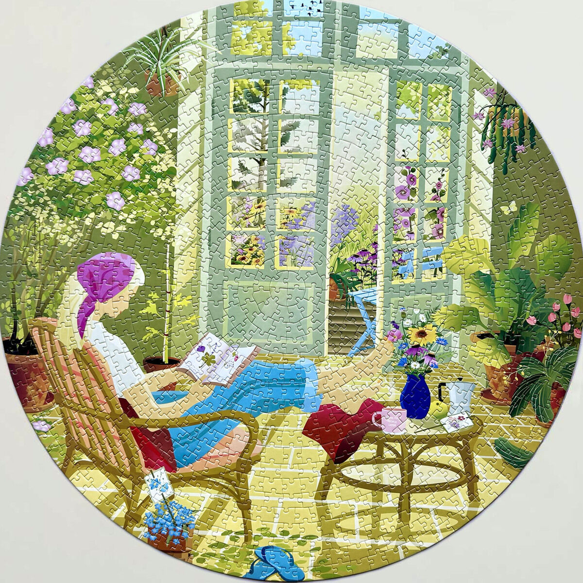 Das runde 1000-Teile-Puzzle »Morning Glow« von ViSSEVASSE zeigt eine entspannte Frau mit Kopftuch, die in einem sonnendurchfluteten Wintergarten sitzt, liest und von vielen Pflanzen in Töpfen und bunten Blumen umgeben ist. Ein kleines Kätzchen schläft auf einer Decke.