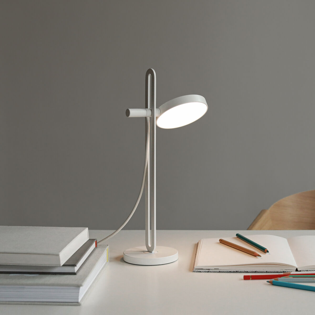 Eine moderne Tischlampe »Echo« steht auf einem weißen Schreibtisch neben einem Stapel Bücher, Stifte und einem offenen Notizbuch. Im Hintergrund ist eine schlichte graue Wand und ein Holzstuhl zu sehen.