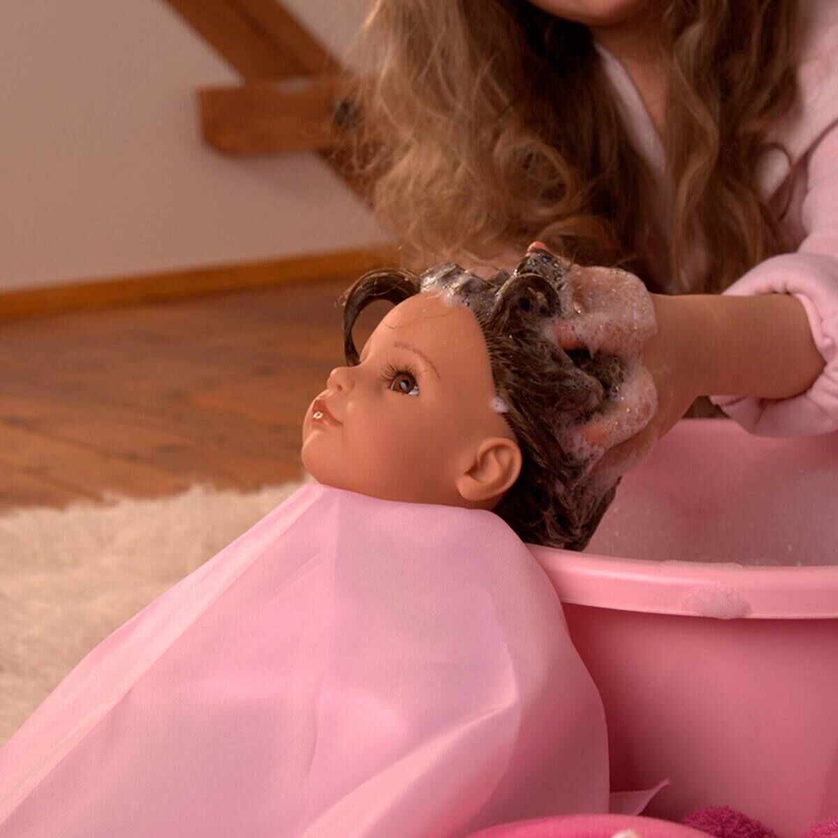 Ein Kind wäscht einer Stehpuppe (Modell „Hannah und ihr Hund Polly“ von Götz Puppen) die Haare in einer rosa Schüssel. Die Szene spielt auf einem Holzboden mit Teppich, die Puppe trägt einen Umhang, der Schaum ist deutlich zu erkennen.