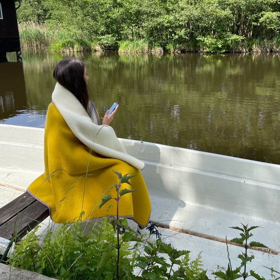 Eine Person sitzt eingewickelt in einer zweifarbigen Decke - gelb und weiß - und blickt auf einen ruhigen See, umgeben von üppigem Grün. Der Himmel ist blau und es scheint ein sonniger Tag zu sein.