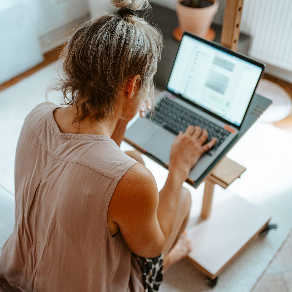 Eine Frau sitzt entspannt auf dem Boden und arbeitet an einem Laptop, der auf dem mobilen Device-Stativ »ERA« aus hellem Massivholz von Frau Caze steht. Das flexible Stativ bietet einen stabilen, eleganten Platz für digitale Geräte, ideal für modernes, kreatives Arbeiten daheim.