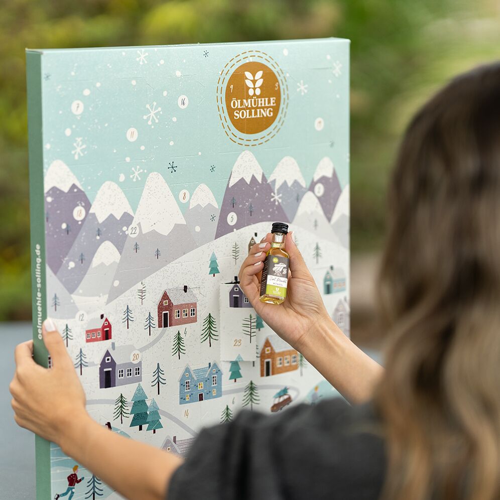 Adventskalender mit winterlicher Dorf- und Berglandschaft, dekoriert mit verschneiten Häusern, Tannenbäumen und Eisläufern; eine Person hält einen kleinen Flascheninhalt vor dem Kalender. Das Produkt eignet sich ideal als Geschenk für die Vorweihnachtszeit und spricht Familien sowie Erwachsene an.
