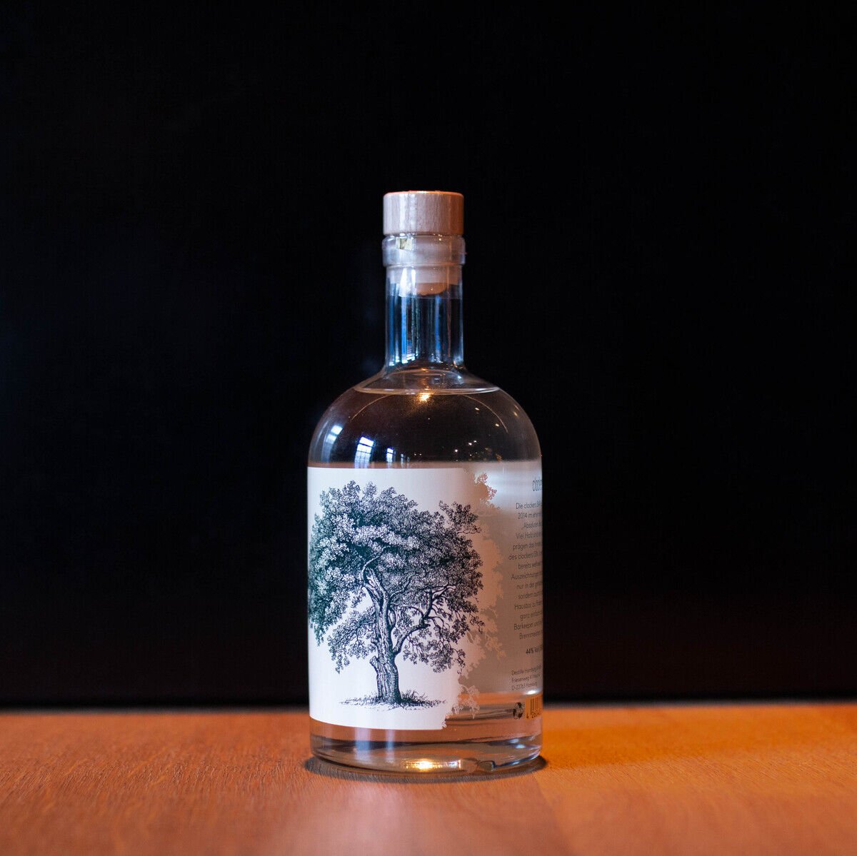 Eine Flasche „clockers Gin St. Pauli“ steht auf einer Holzoberfläche vor einem schwarzen Hintergrund. Die Flasche ist klar und trägt ein Etikett mit einem detaillierten Baumdesign.