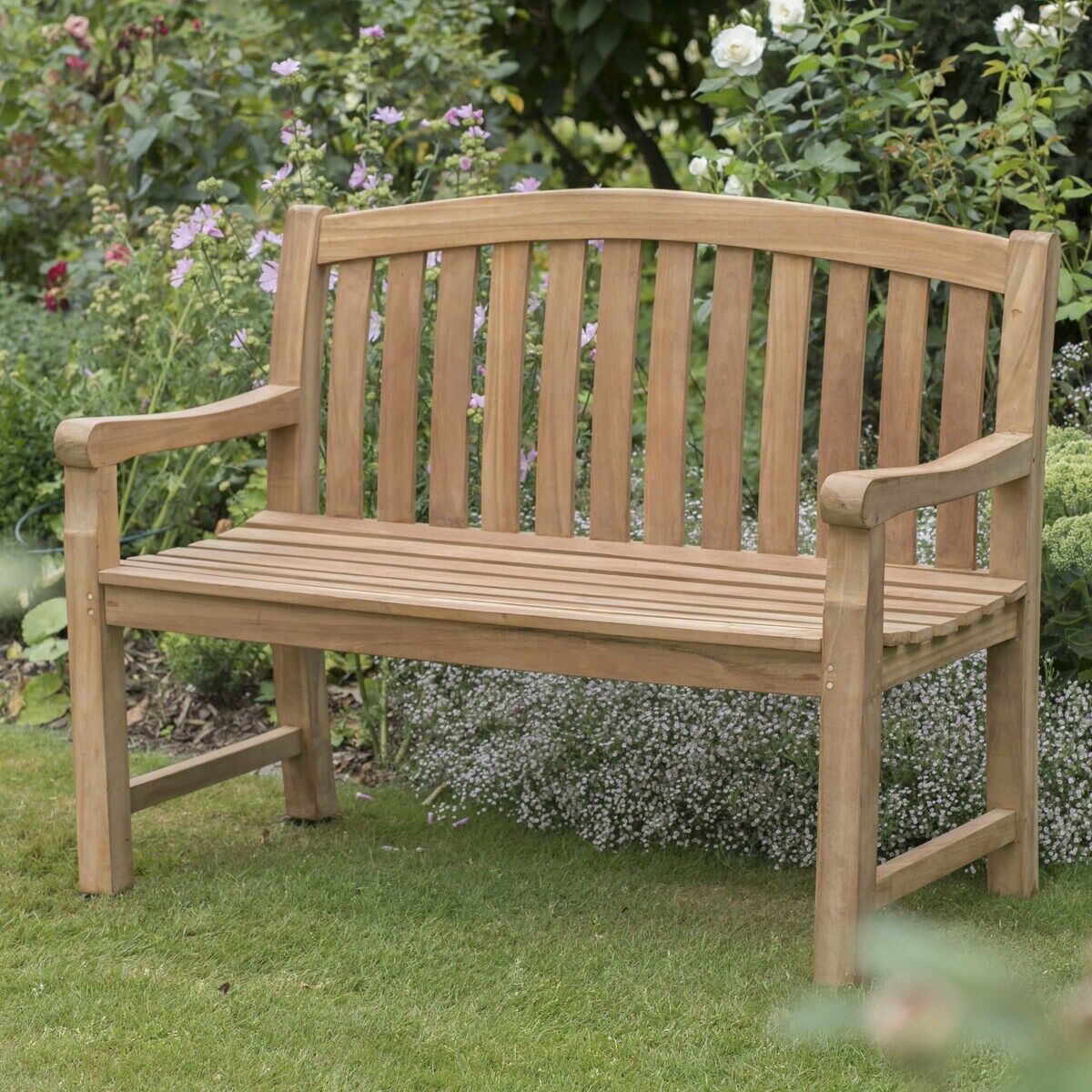 Holz-Gartenbank »Swindon« von bene living steht auf einem gepflegten Rasen, umgeben von blühenden Blumen und dichtem grünen Laub. Die Bank besteht aus hellem Teakholz mit geschwungener Rückenlehne und Armlehnen und wirkt hochwertig verarbeitet. Ruhige, einladende Gartenszene.