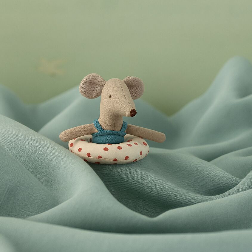 Ein kleines Stofftier in Form einer Maus sitzt auf einer gepunkteten Luftmatratze vor einem pastellblauen Hintergrund. Die Maus trägt einen blauen Badeanzug. Das Bild vermittelt eine sommerliche, verspielte Stimmung und eignet sich zur Darstellung von Badespaß und Strandmotiven für Kinder.