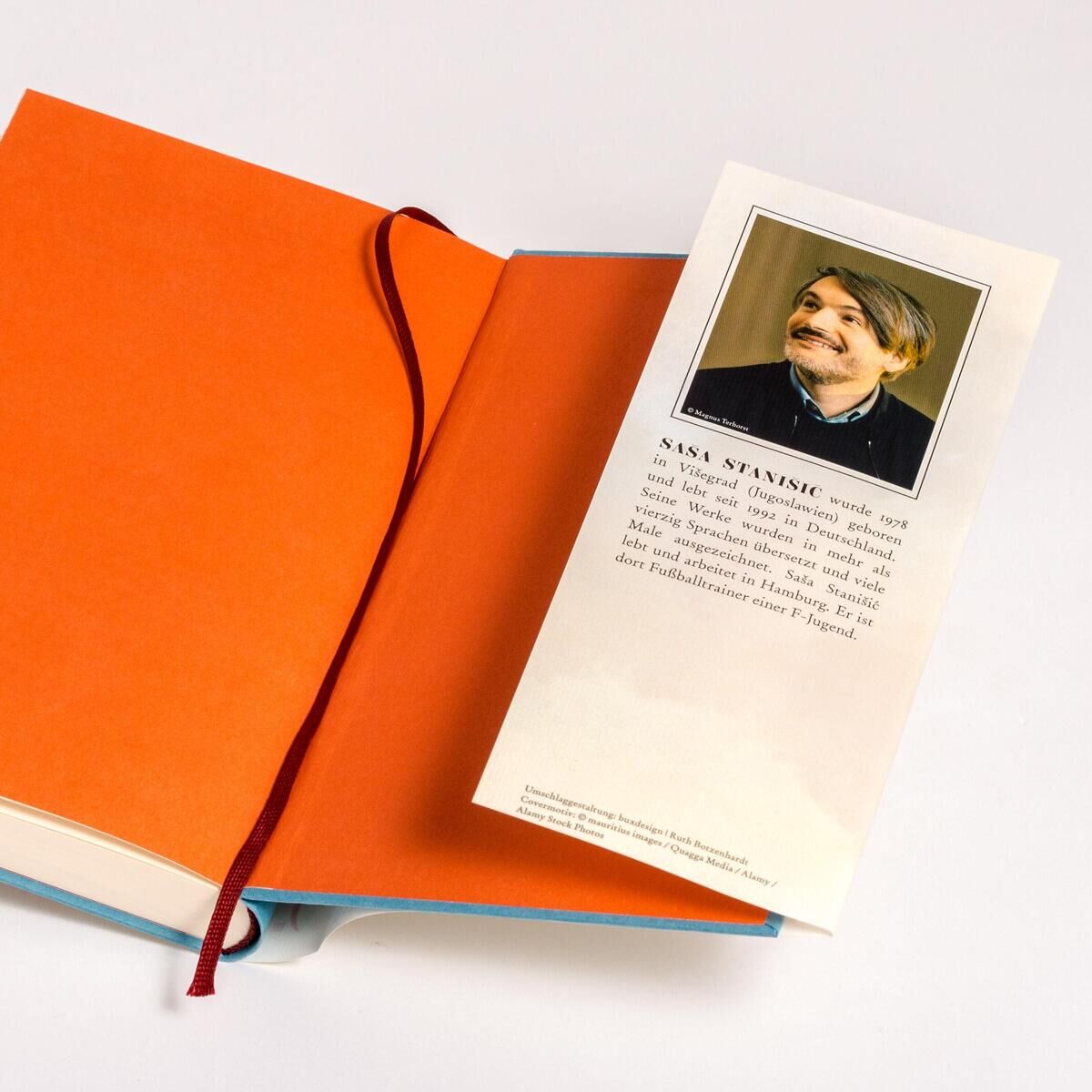 Ein geöffnetes Buch mit orangefarbenen Seiten. Ein rotes Lesezeichen ist in der Mitte platziert. Oben rechts ist ein Platzhalter für ein Foto mit einer Beschreibung darunter.