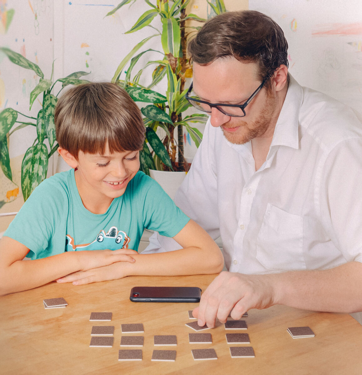 Ein Vater und sein Junge sitzen an einem Holztisch und spielen ein Gedächtnisspiel. Der Vater trägt ein weißes Hemd und schwarze Brille, während der Junge ein grünes T-Shirt trägt. Im Hintergrund sind grüne Pflanzen zu sehen. Auf dem Tisch liegen Karten und ein Smartphone.