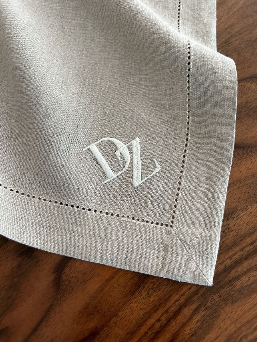 Eine naturfarbene Leinenserviette mit einem aufgestickten weißen Monogramm in der Ecke, auf einem dunklen Holztisch platziert.