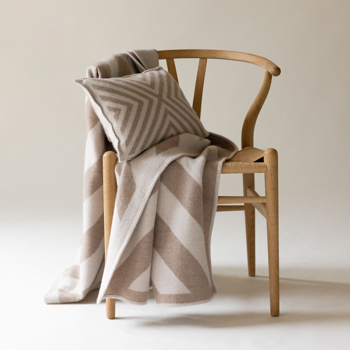 Ein Kaschmir Kissenbezug namens 'Pontresina', platziert auf einem hölzernen Stuhl, kombiniert mit einer weichen Kaschmirdecke in beige und weißen Farben, mit geometrischen Mustern in einem schlichten, eleganten Raum.