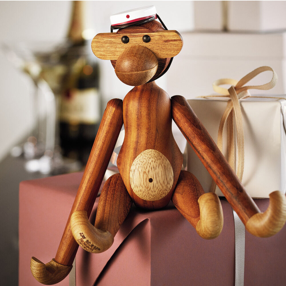 Auf dem Bild sitzt die mittlere Kay Bojesen Holzfigur »Affe« aus Teak- und Limbaholz mit betontem Gesichtsausdruck und rotem Mützchen auf zwei liebevoll verpackten Geschenken. Im Hintergrund ein Weinglas und eine Flasche, Ambiente festlich und einladend.