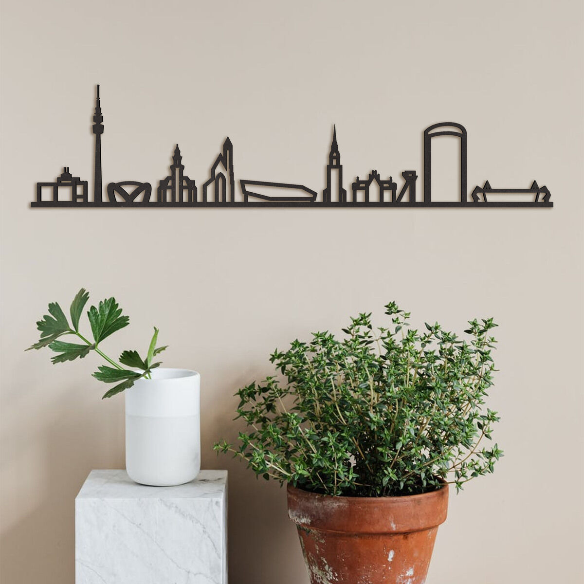 Eine dekorative Skyline von Dortmund aus Holz, an einer neutralen Wand hängend mit zwei Zimmerpflanzen in Pflanzgefäßen davor.