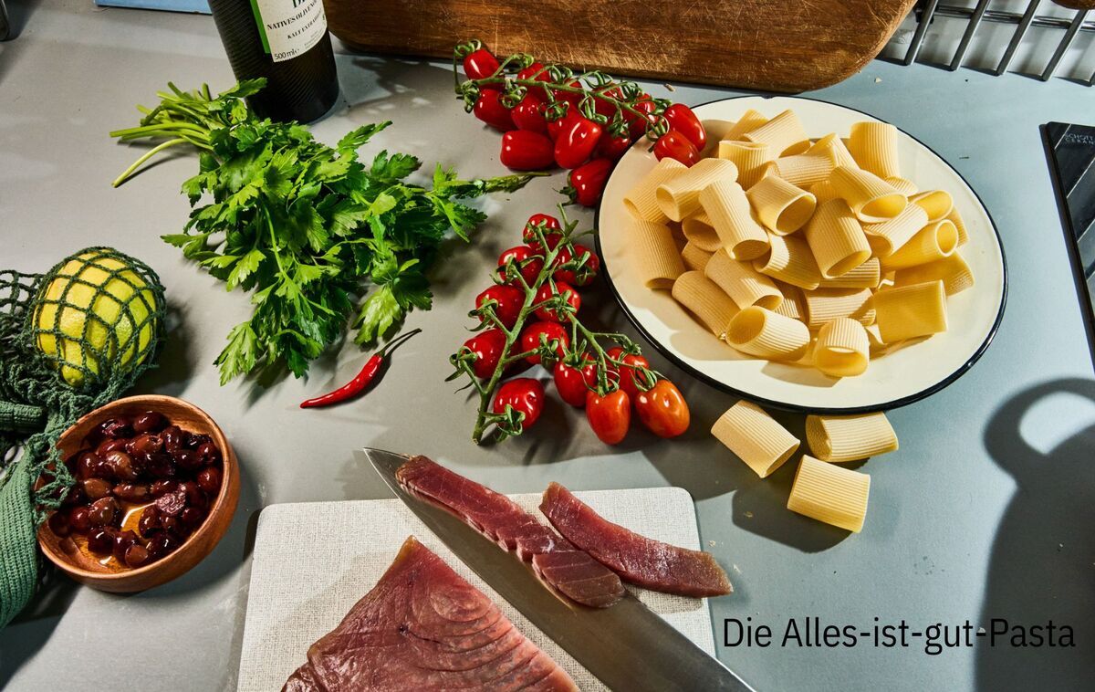 Auf einer Küchentheke liegen Zutaten für ein mediterranes Gericht: ein Bund Petersilie, Kirschtomaten an der Rebe, eine Schüssel mit großen Nudelrohren, ein Stück roher Thunfisch, rote Chilischoten und ein Netz mit Zitrone. Eine Flasche Wein steht im Hintergrund.