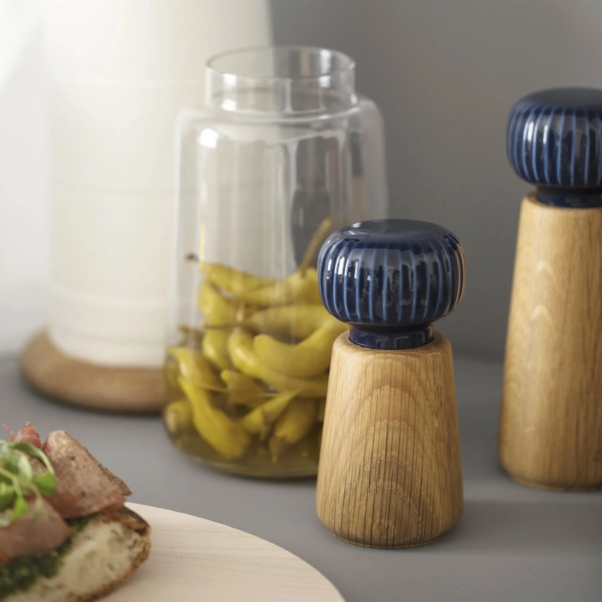 Zwei stilvolle Gewürzmühlen der Serie Hammershøi mit dunkelblauen Keramikköpfen und Holzsockel stehen auf einem hellen Tisch. Dahinter befinden sich ein Glas mit eingelegten Peperoni und ein weiterer Behälter. Im Vordergrund ist ein belegtes Brot angeschnitten zu sehen. Das Arrangement betont modernes skandinavisches Design und warme Kücheneinrichtung.