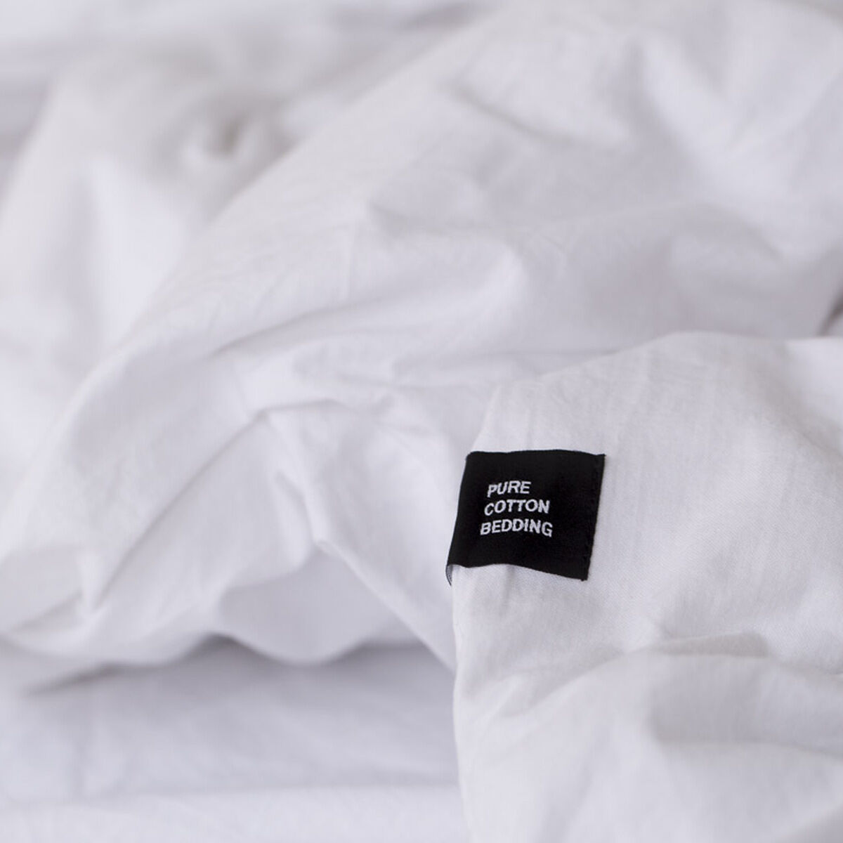 Nahaufnahme von weißer Bettwäsche »Pure« aus 100% Baumwolle mit einem schwarzen Etikett mit der Aufschrift 'PURE COTTON BEDDING'.