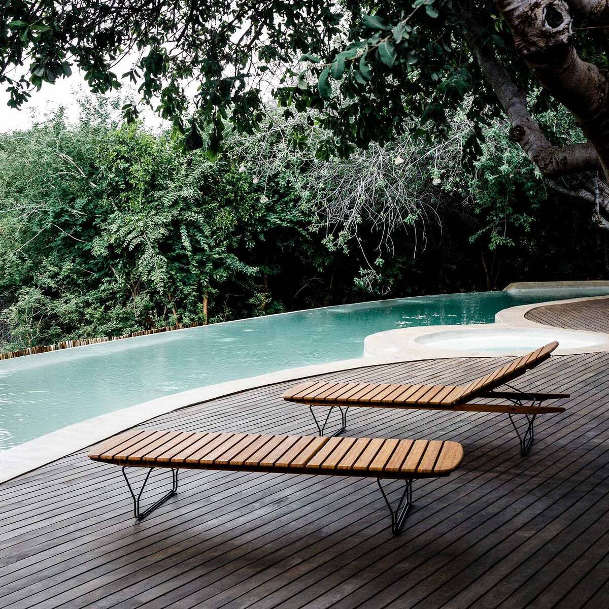 Zwei Holzsonnenliegen stehen auf einer Holzterrasse neben einem Swimmingpool, umgeben von dichter, grüner Vegetation und Bäumen.