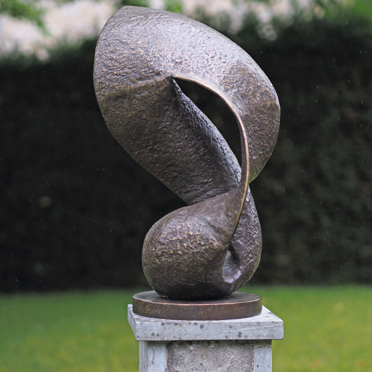 Bronzene Gartenskulptur in Möbiusband-Form auf rechteckigem Steinsockel, vor grünem, unscharfen Hintergrund. Die Oberfläche ist strukturiert, weich glänzend und wirkt durch Drehungen und Schwünge dynamisch-modern. Maße etwa 55 cm hoch.