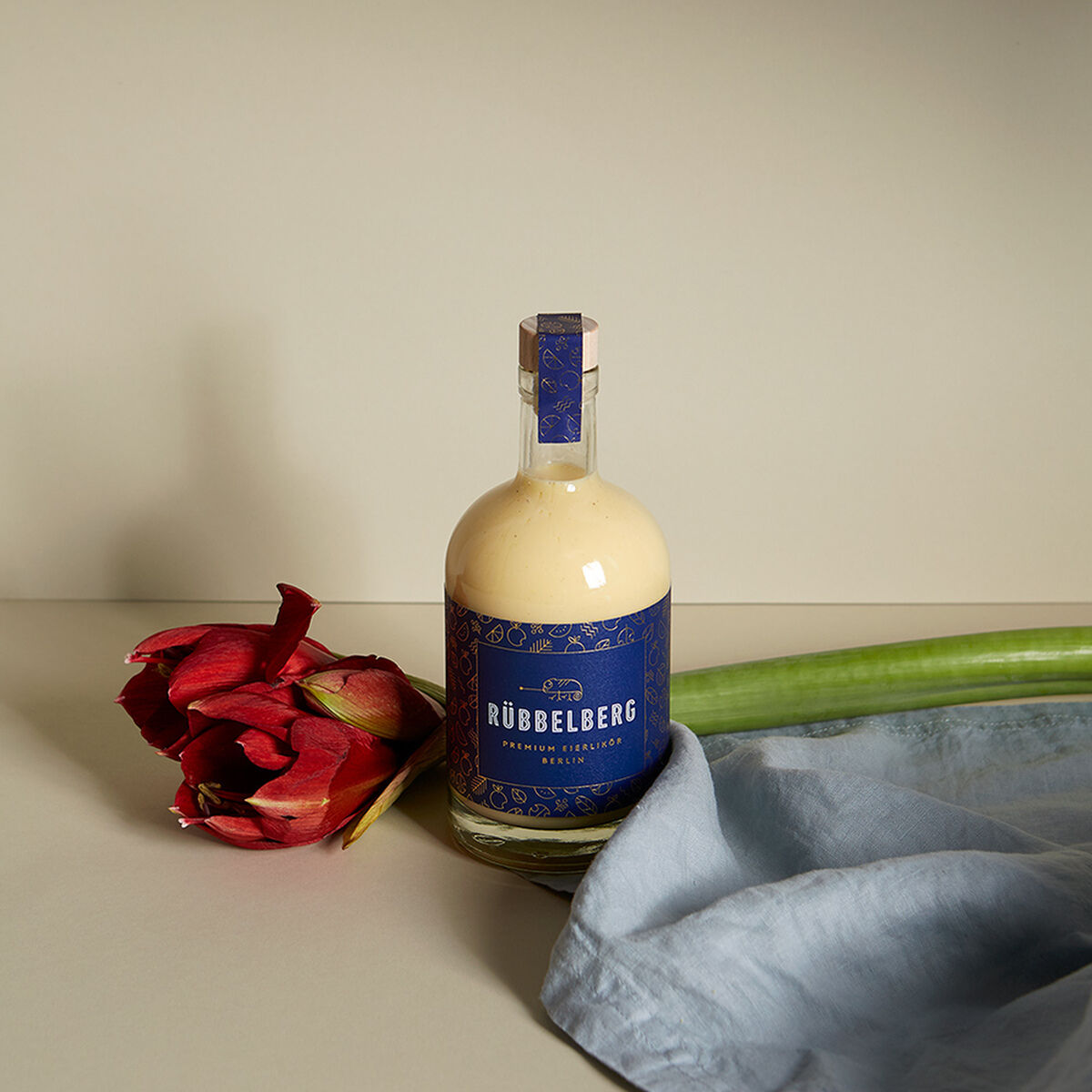 Eine Flasche Premium Eierlikör von Rübbelberg steht auf einem Tisch neben einer roten Blume und einem blauen Stoff. Die Flasche hat ein blaues Etikett und ist in einer neutralen Umgebung positioniert.
