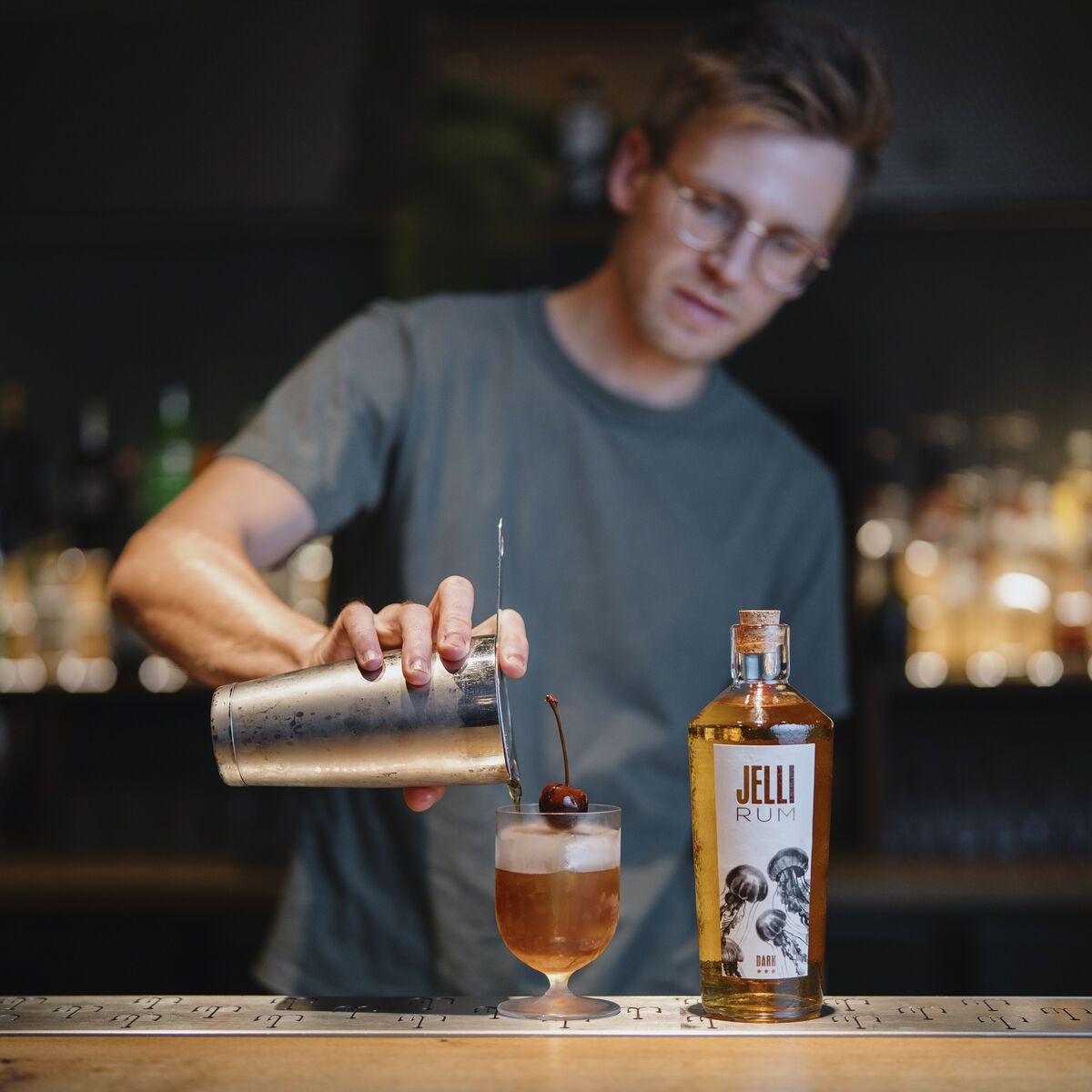Ein junger Mann in legerem T-Shirt bereitet an einer stilvollen Bar einen Cocktail zu, indem er einen Drink mit Shaker in ein Glas eingießt. Neben dem Glas steht eine Flasche JELLI Rum, Dark. Auf dem Cocktail liegt eine Kirsche als Garnitur. Im Hintergrund ist eine Bar mit verschiedenen Flaschen zu sehen.