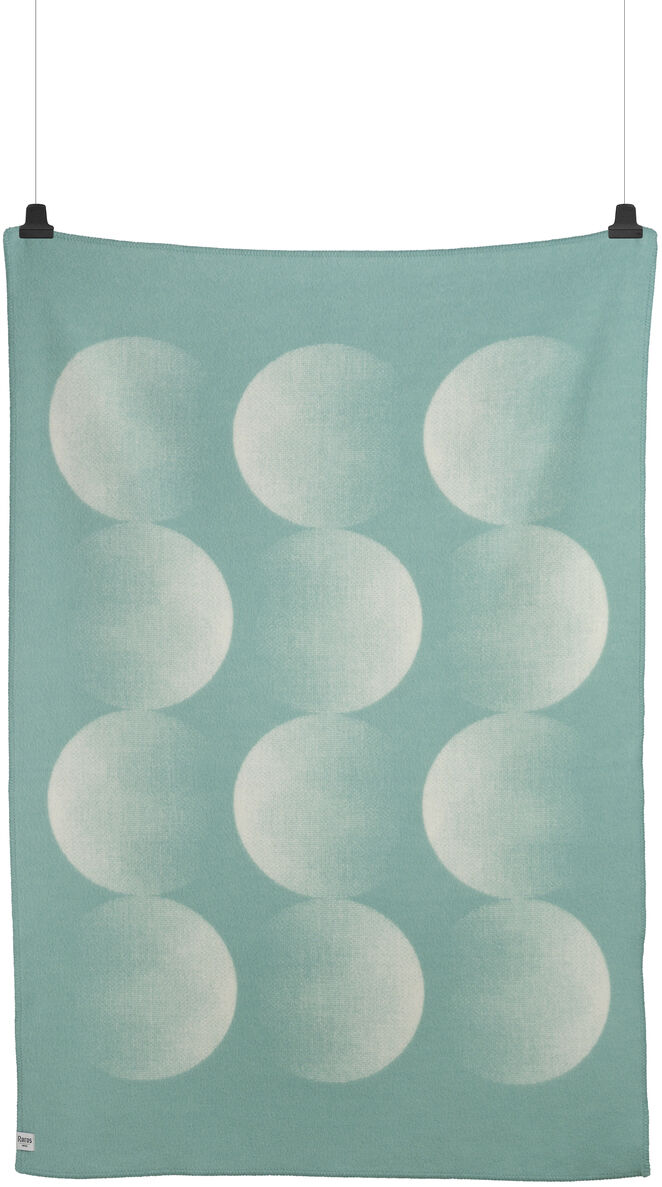 Eine Lammwolldecke namens MOON in Grün mit einem weißen Muster, das an Mondphasen erinnert. Die Decke hängt an zwei schwarzen Clips.