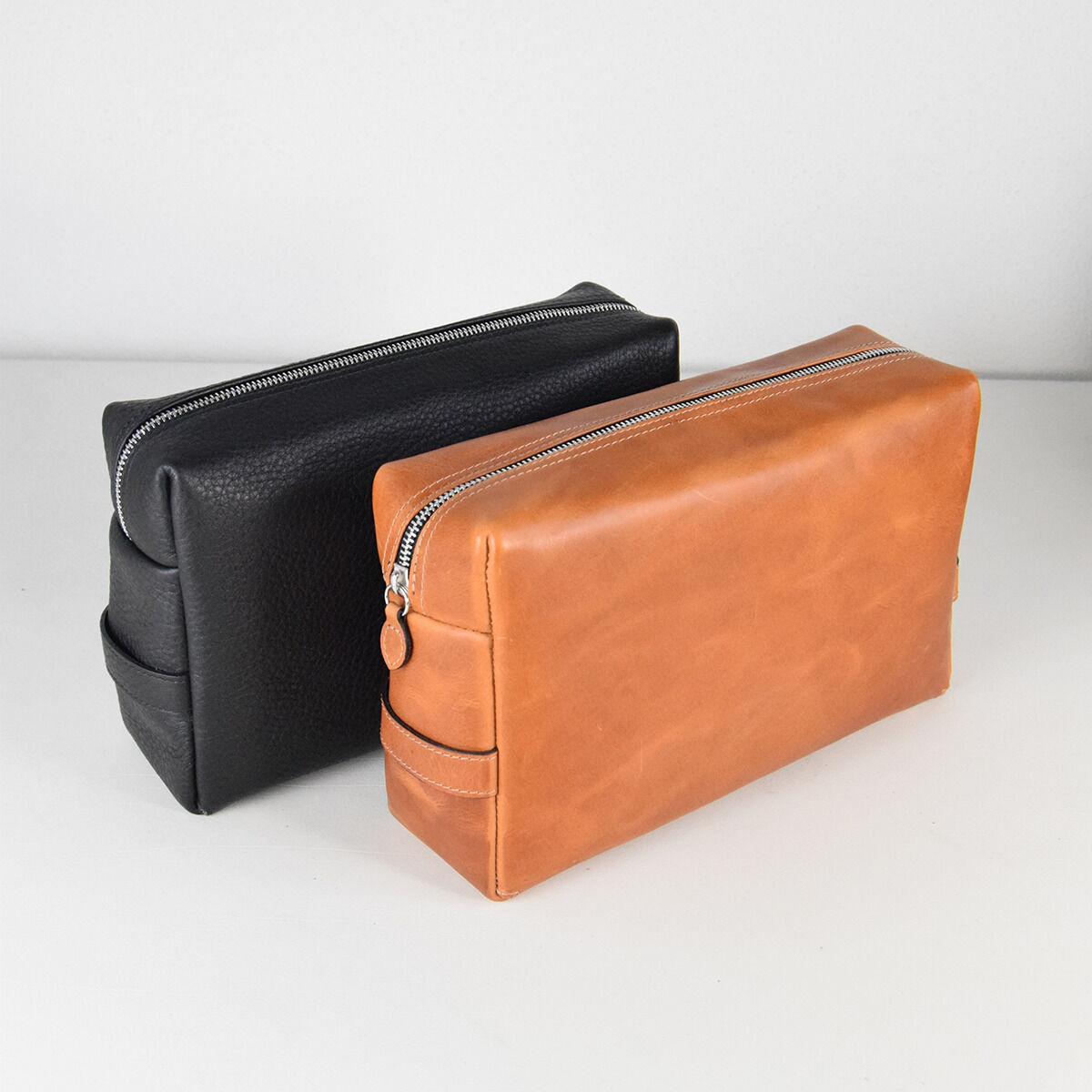 Zwei Kulturtaschen aus Leder, eine in Schwarz und eine in Braun, auf einer weißen Oberfläche. Sie sind rechteckig, mit Reißverschlüssen oben und einem minimalistischen Design.