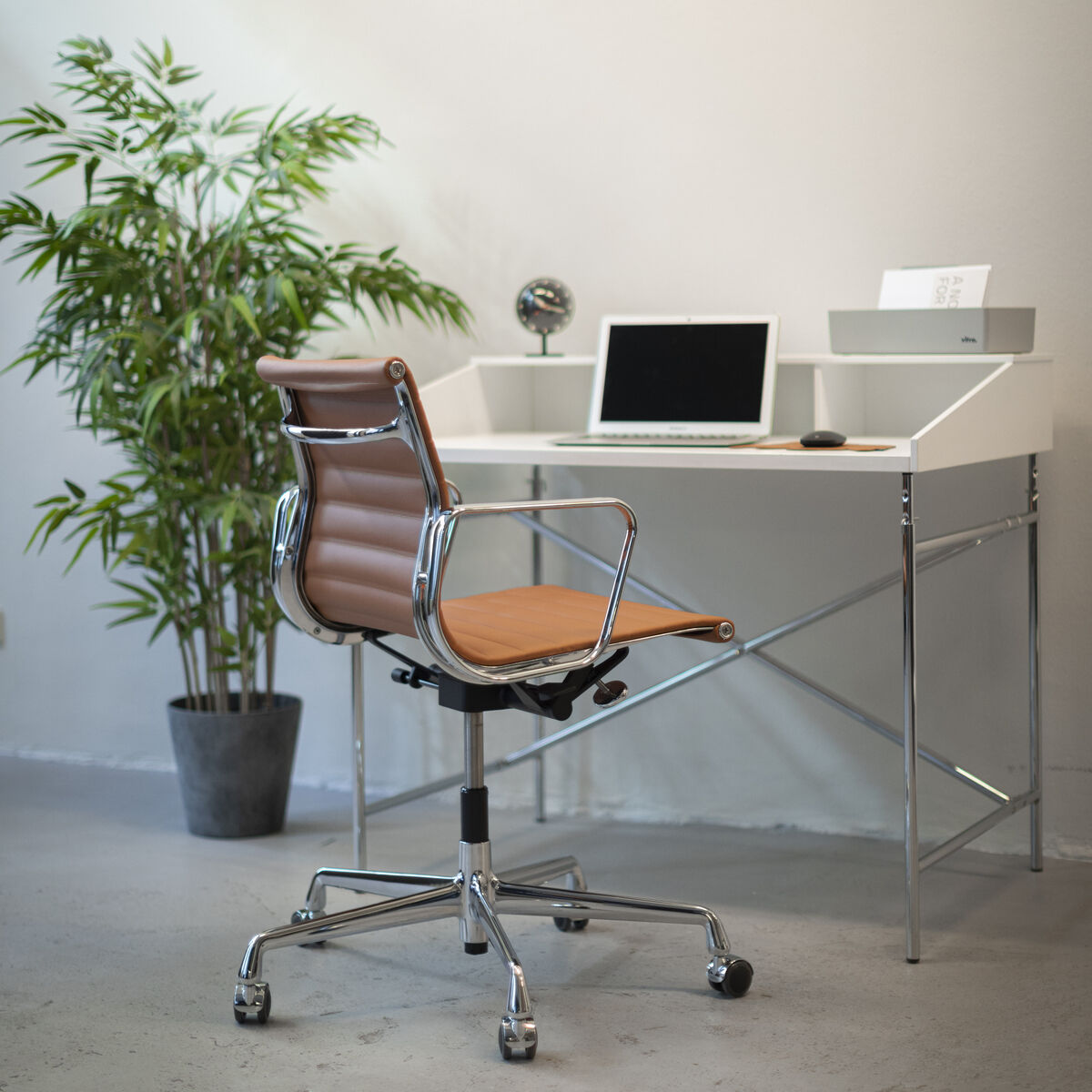 Design-Szene mit Schreibtischstuhl »Vitra Alu Chair EA 118« in cognacfarbenem Leder und verchromtem Aluminium, vor weißem Schreibtisch mit Laptop und Deko. Daneben steht eine große grüne Pflanze in grauem Topf auf hellem Boden, Hintergrund helle Wand.