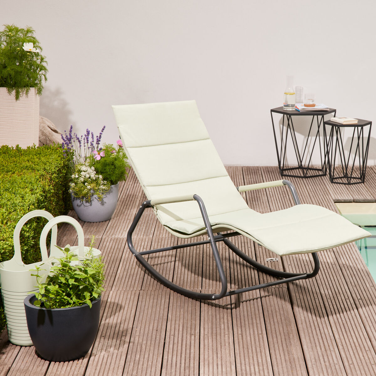 Eine moderne, cremeweiße Swingliege »Aliano« steht auf einer Holzterrasse neben einem Pool, umgeben von grünen Pflanzen und kleinen Beistelltischen mit Getränken. Badetuch und Gießkanne ergänzen die sommerliche Gartenszene.