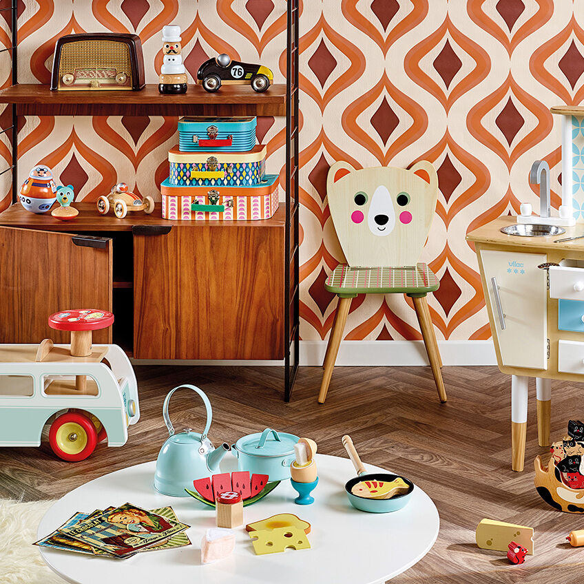 Buntes Kinderzimmer mit verschiedenen Spielzeugen, darunter ein Minibus aus Holz, eine Puppenküche und ein Stapelspielzeug. Der Raum ist fröhlich und farbenfroh eingerichtet mit Retro-Tapetenmuster und Holzmöbeln. Einige Pflanzen und Dekoartikel verleihen dem Zimmer eine gemütliche Atmosphäre.