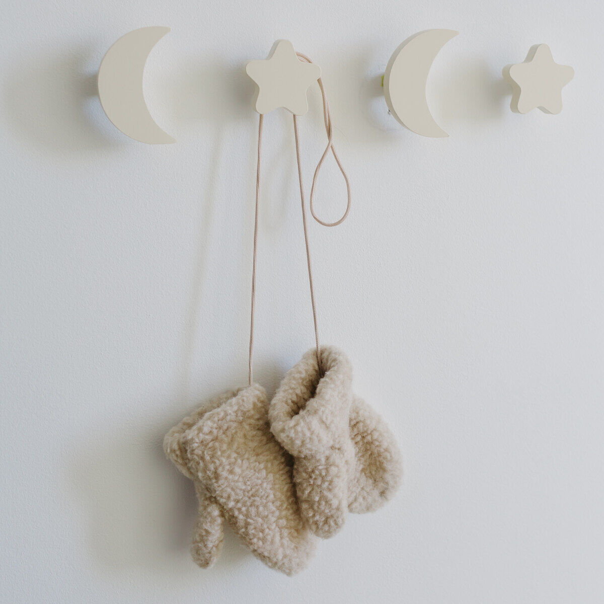 Eine Wandhakenleiste in Form von Monden und Sternen, an der beigefarbene Handschuhe hängen. Die Haken und die Wand sind in warmen, neutralen Farben gehalten.
