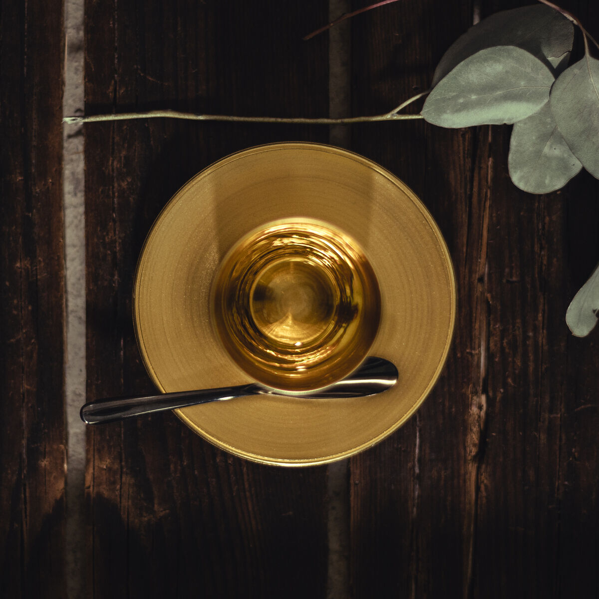 Ein goldgelber Espressobecher auf passendem Unterteller mit einem glänzenden Löffel darauf, arrangiert auf dunklem Holztisch neben dekorativen graugrünen Zweigen. Die Szene vermittelt einen modernen und gemütlichen Kaffeehausstil.