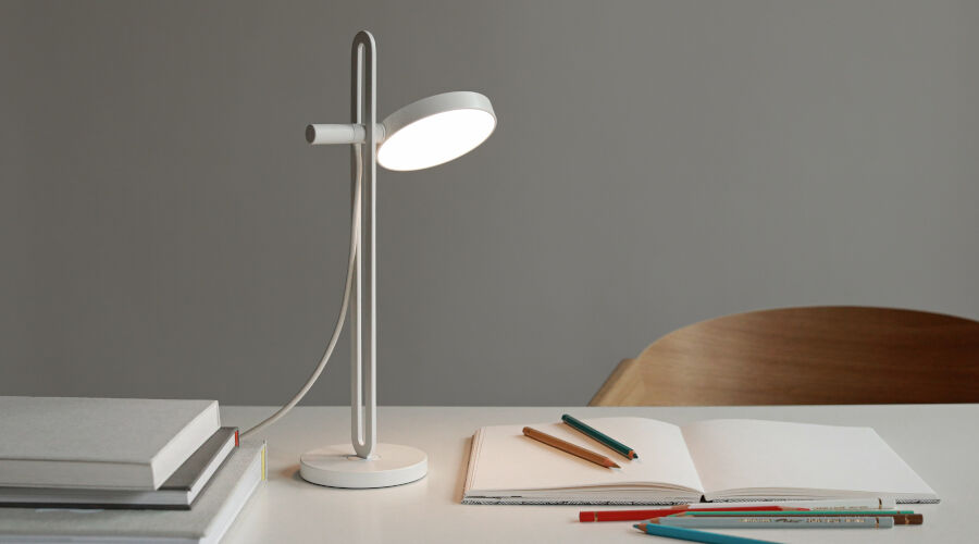 Moderne weiße Schreibtischlampe mit rundem Lampenschirm auf hellem Schreibtisch neben geöffnetem Notizbuch und Buntstiften
