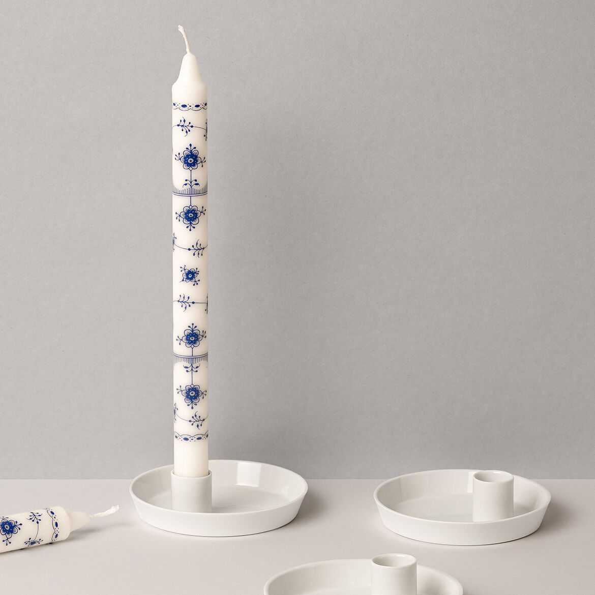 Ein Porzellan-Kerzenhalter mit schlichter, moderner Form, der eine weiße Kerze mit blauen, handgemalten Verzierungen hält, auf einem neutralen Hintergrund präsentiert.