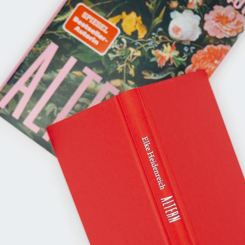 Das Bild zeigt das Buch 'Altern' von Elke Heidenreich. Es liegt auf einem weiteren Buch, das mit bunten Blumen bedruckt ist. Das vordere Buch ist leuchtend rot mit weißer Schrift auf dem Rücken.
