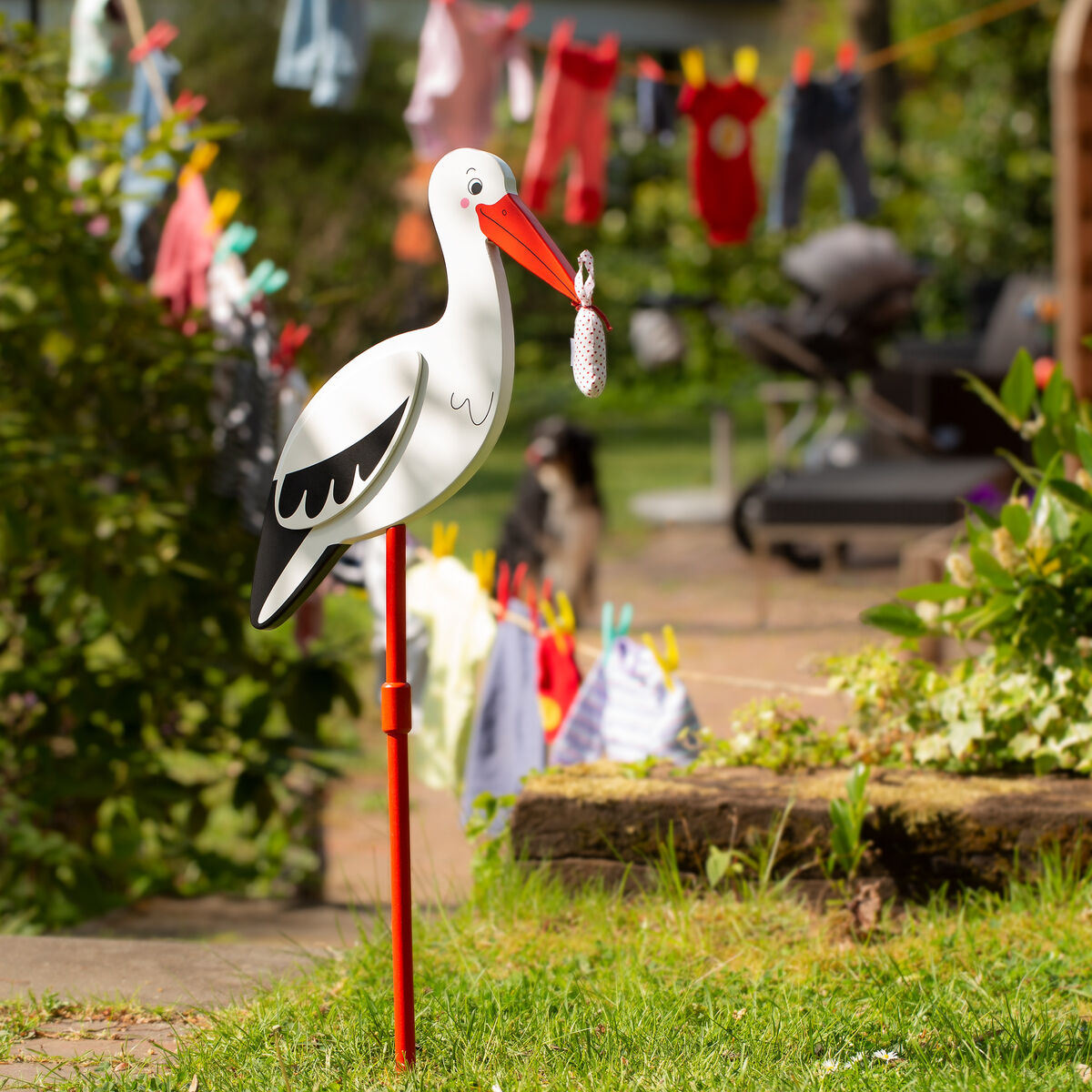 Ein Stecker in Form eines Storchs, der ein Babytuch im Schnabel hält, steht in einem Garten. Im Hintergrund hängt bunte Babykleidung an einer Wäscheleine.