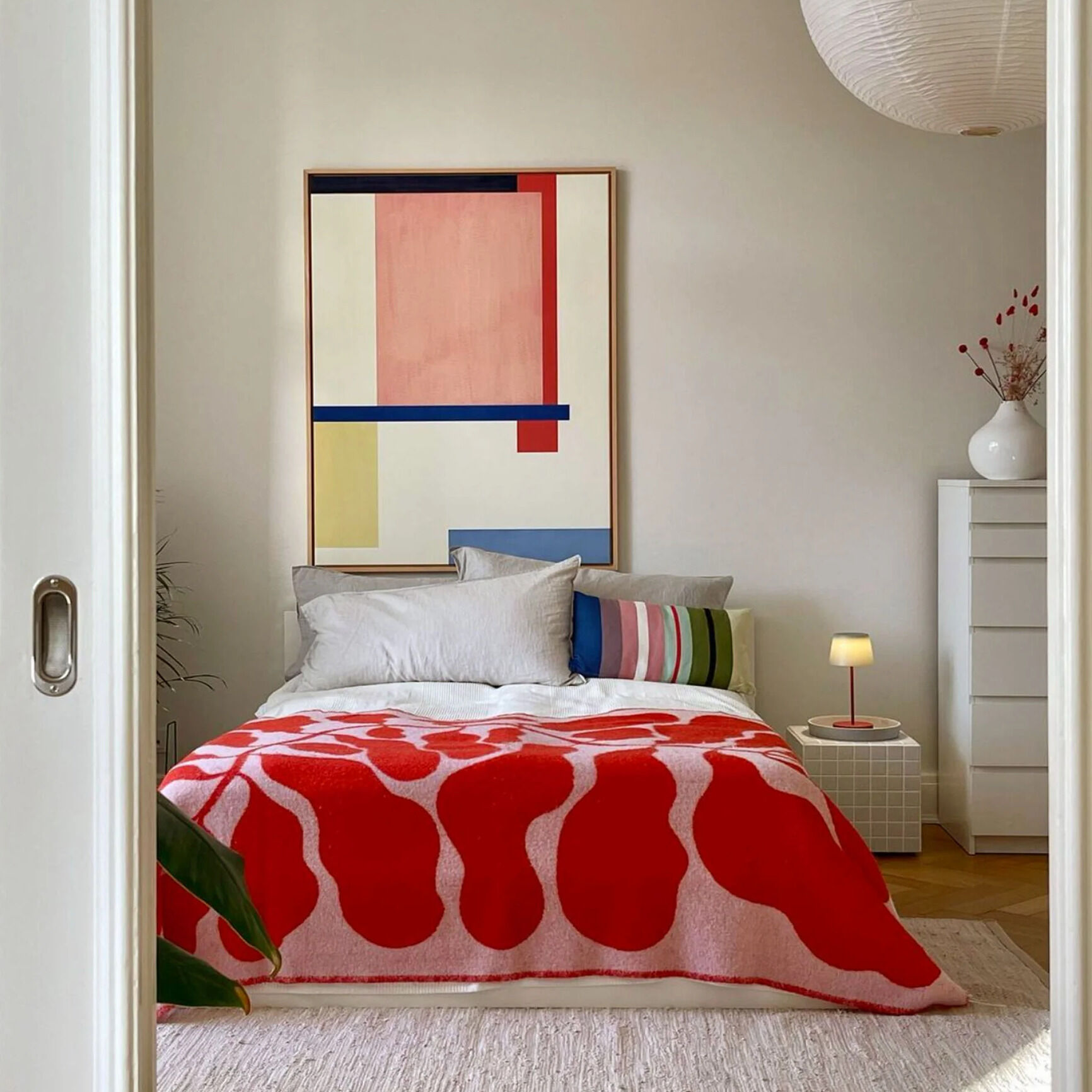 Geräumiges Schlafzimmer mit modernem Leinwandbild »Fokke No 02« an der Wand, das geometrische, farbige Formen zeigt. Im Vordergrund ist ein Bett mit roter und weißer Decke, daneben stehen ein weißer Nachttisch, eine kleine Lampe und eine weiße Kommode mit Vase und getrockneten Zweigen.