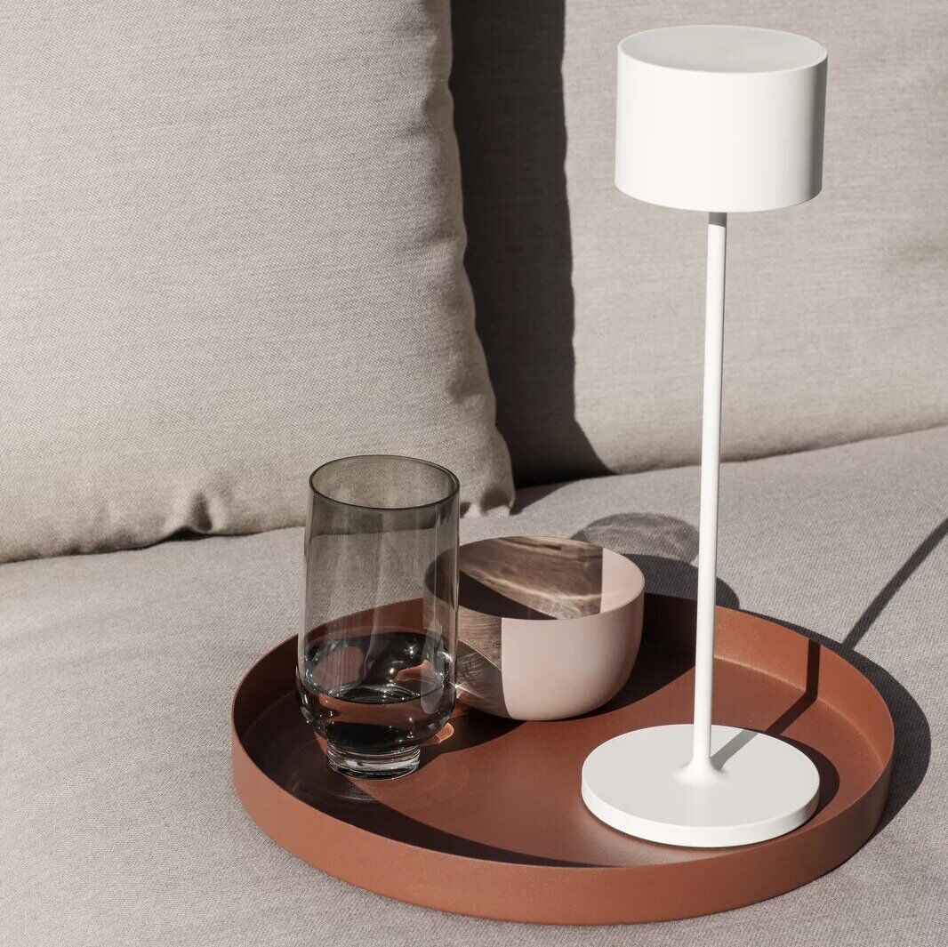 Eine mobile LED-Tischleuchte namens »FAROL« steht auf einem braunen Tablett neben einem Glas und einer kleinen Schale auf einem beigen Sofa.