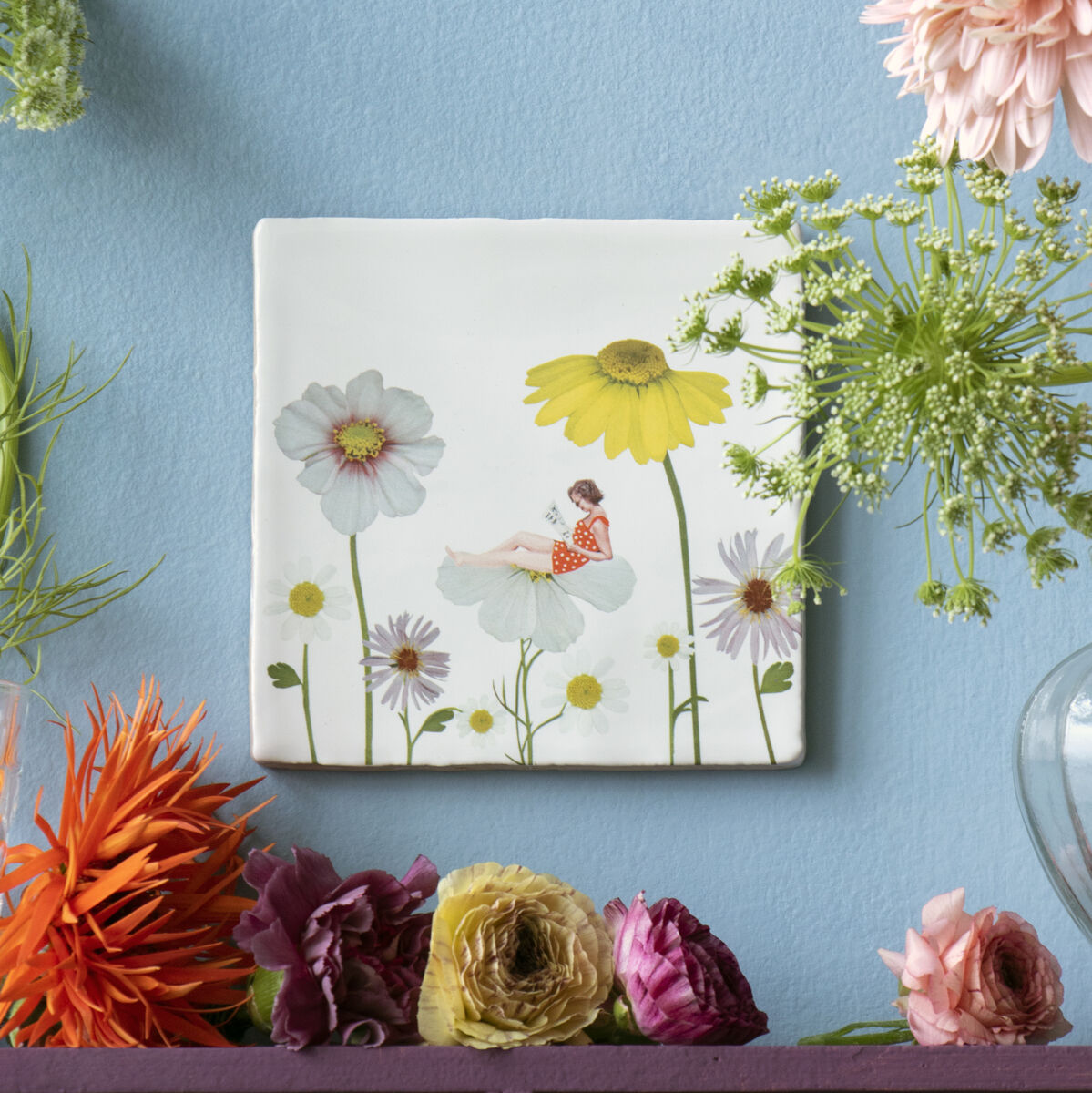 Keramikfliese mit Blumendesign an einer blauen Wand. Vor der Fliese befinden sich Blumen in verschiedenen Farben in Glasvasen und auf einem lila Regalbrett.