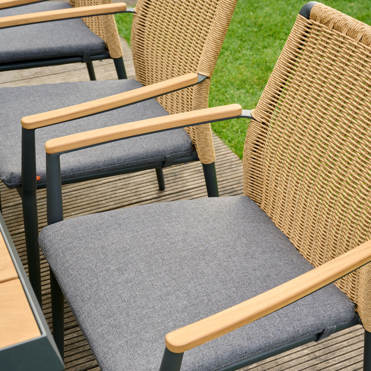 Ein Stapelsessel mit einem Metallrahmen, Armlehnen aus Holz und einer Sitzfläche in Rattan-Optik steht auf einer Terrasse mit Holzboden, umgeben von grünem Rasen.