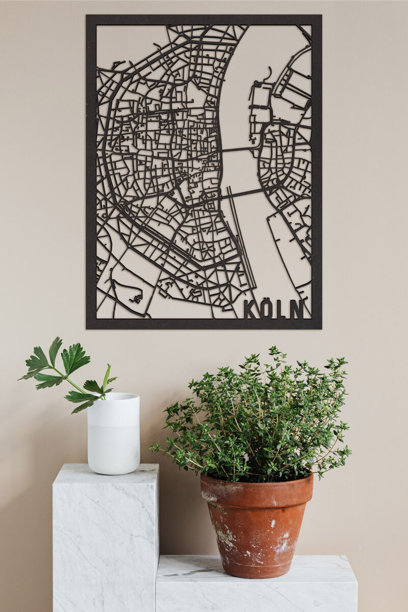 Ein detaillierter Holzstadtplan von Köln, der als Wanddekoration dient. Der Stadtplan ist schwarz und zeigt die Straßen und Flüsse von Köln. Er hängt an einer beigen Wand, darunter eine weiße Kerze auf einem weißen Marmornpodest und ein grüner Pflanzenstrauch in einem Terrakotta-Topf.