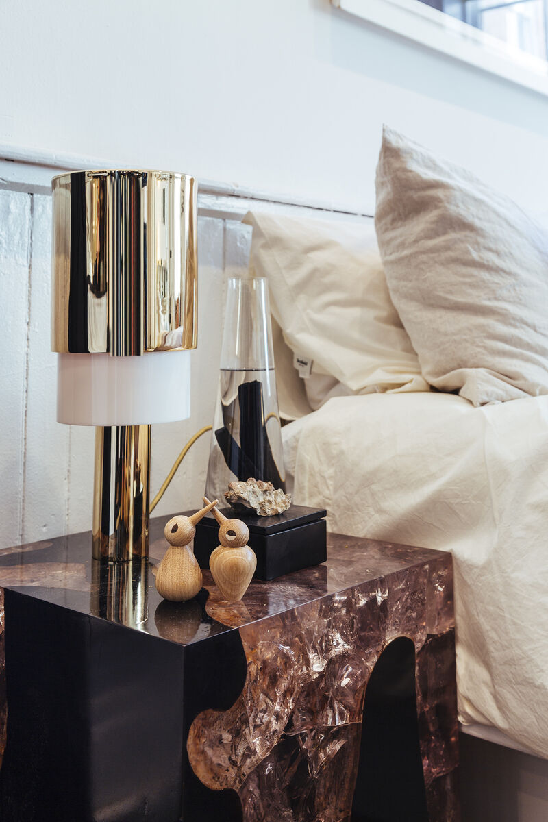 Holzfiguren in Vogelgestalt auf einem Nachttisch aus dunklem Holz neben einer goldenen Lampe und einem Bett mit beiger Bettwäsche im skandinavischen Design.