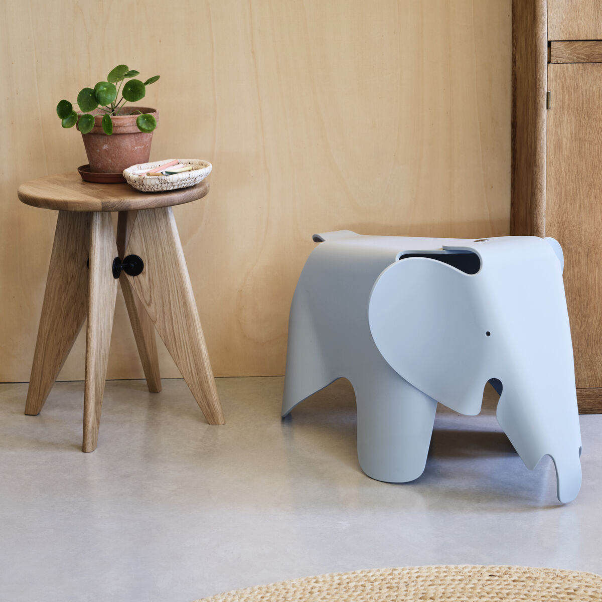 Kinderhocker in Form eines Elefanten, namens »Eames Elephant«, in Hellgrau neben einem runden Holztisch mit Pflanze darauf. Die Szene ist in einem eleganten, modernen Innenraum mit Holz- und Pastelltönen.