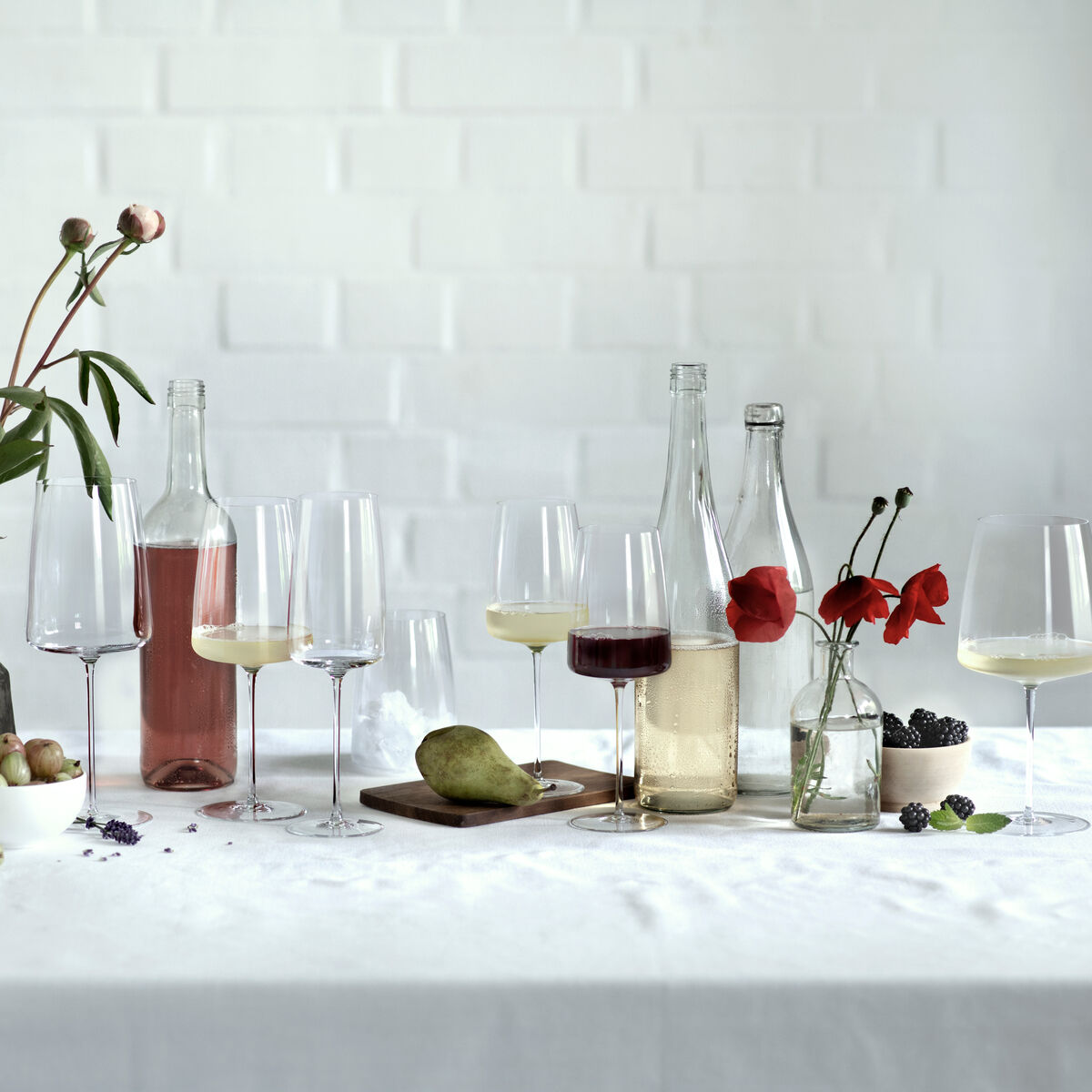 Ein stilvoll dekorierter Tisch mit einem Rotwein-Zweiterset »Enoteca«, bestehend aus mehreren Weingläsern, Weinflaschen und dekorativen Elementen wie Blumen und Früchten. Hauptfarben sind Weiß, Rot und Grün.