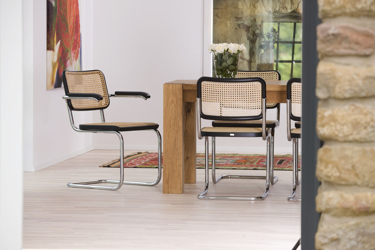 Das Bild zeigt zwei S 32 Freischwinger-Stühle von Thonet in einem modernen Esszimmer mit einem Holztisch und Rattangeflecht. Die Stühle haben ein schwarzes Metallgestell und beige Sitzflächen. Ein bunter Teppich liegt auf heller Holzboden und Blumen befinden sich auf dem Tisch.