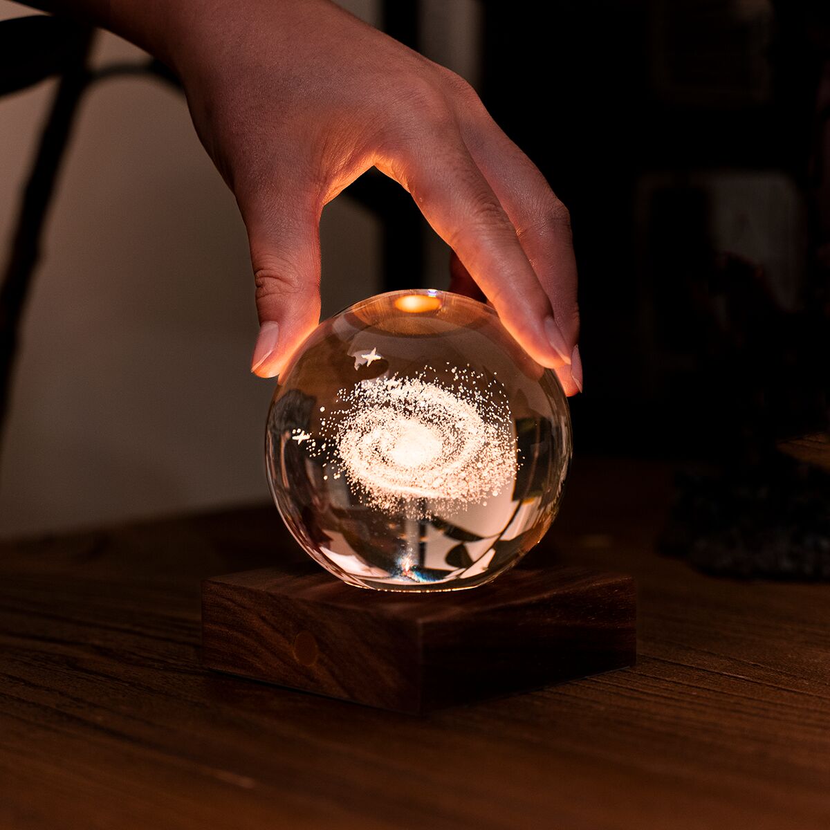 Tischlampe Amber Light mit einer runden Glaskugel und Holzsockel, die im Inneren eine funkelnde Galaxie zeigt. Die Lampe strahlt warmes, bernsteinfarbenes Licht ab und steht auf einem Holztisch in einem gemütlich eingerichteten Raum.